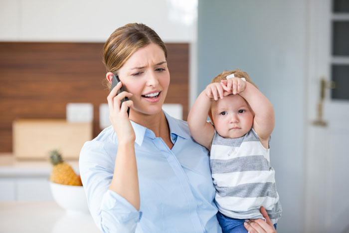 Woman on phone holding baby, showing frustration in a modern kitchen, highlighting travel needs and friendship challenges.