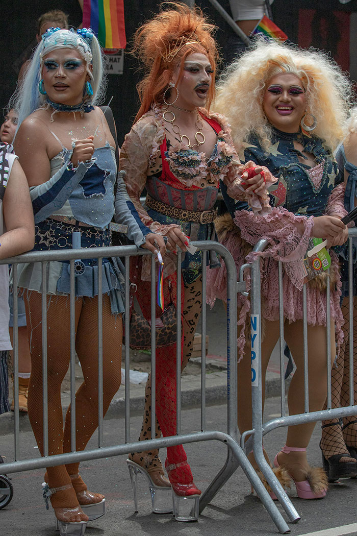 Two amazing drag queens in vibrant outfits stand behind barricades at a lively outdoor event celebration.