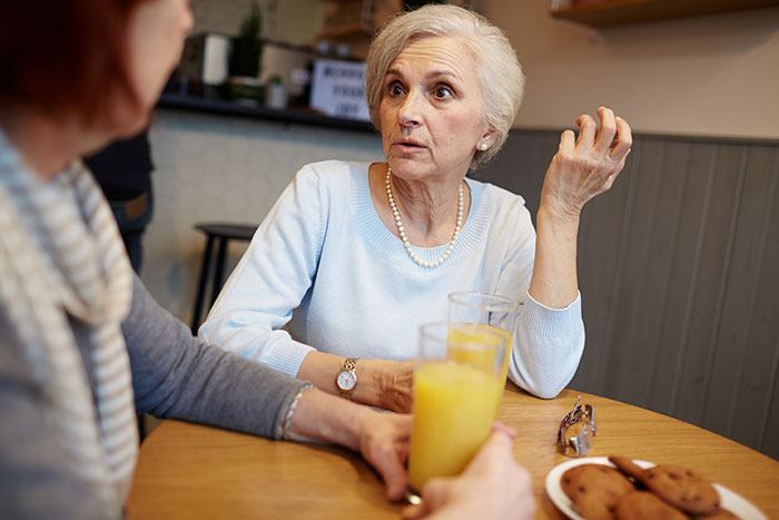 Older woman appearing upset while talking to another person at a table with drinks and cookies, mother-in-law wedding conflict concept.