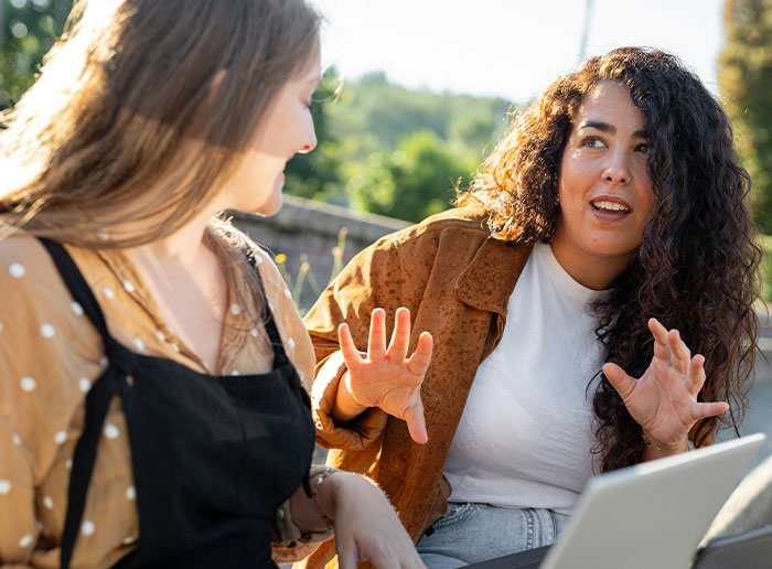 Teen friends having a serious conversation outdoors during the day, one looking confused and the other explaining something.
