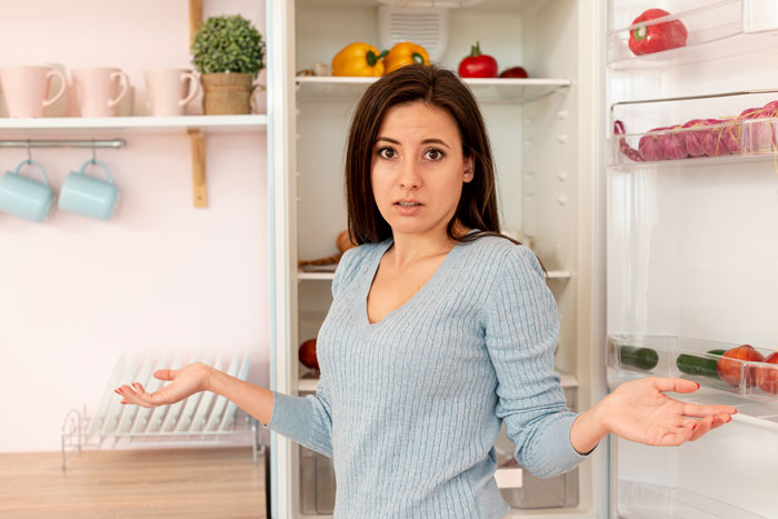 Woman standing confused in front of an open fridge, reacting to leftover pasta theft and considering a mini fridge purchase.