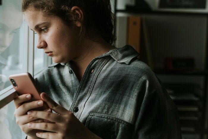 Young woman looking concerned and holding phone near window, highlighting friend stop messaging friendship feelings.