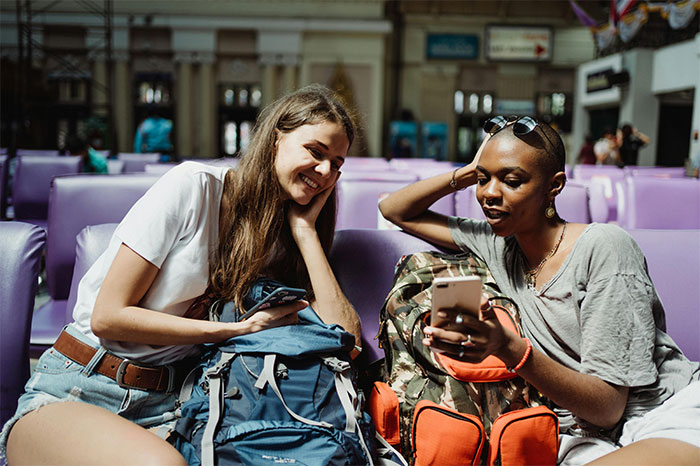 Two women with backpacks sit in a station, one showing the other something on her phone, highlighting travel and friendship moments. Two women with backpacks sit in a station, one showing the other something on her phone, highlighting travel and friendship moments.