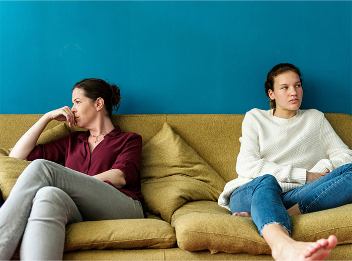 Two women sitting apart on a couch, looking upset, illustrating a woman telling friends she is not a hotel after trips. Two women sitting apart on a couch, looking upset, illustrating a woman telling friends she is not a hotel after trips.
