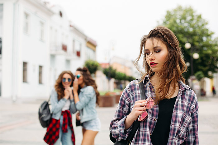 Young woman looking upset and distant while two friends whisper and laugh in the background during a trip outdoors Young woman looking upset and distant while two friends whisper and laugh in the background during a trip outdoors