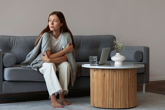Woman sitting on couch wrapped in blanket, looking upset and reflective after catching husband cheating with her sister.