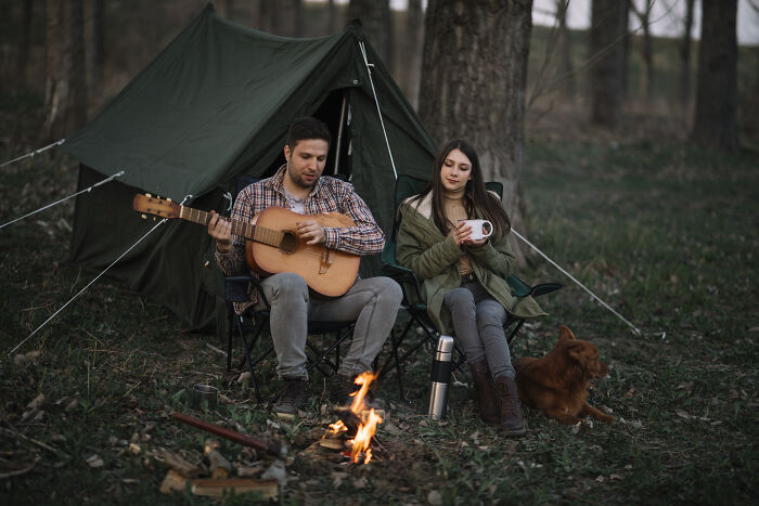 Couple camping in forest with guitar and campfire, enjoying a peaceful outdoor moment with their dog nearby.