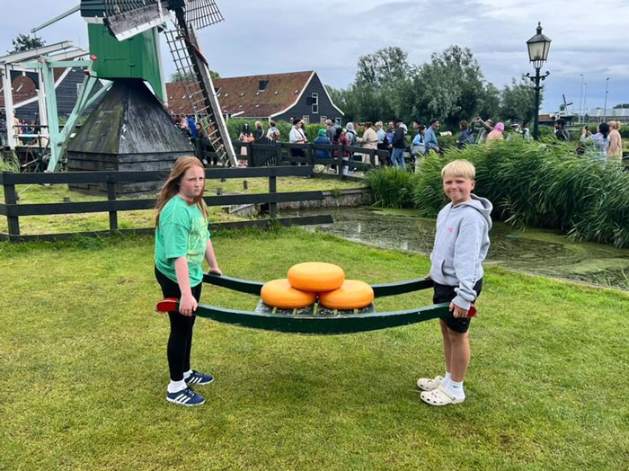 Two kids holding a wooden cheese carrier with large cheese wheels, posing for funny vacation pics outdoors by a windmill.