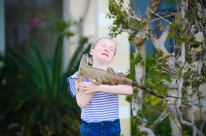 Young girl struggling to hold large iguana in funny vacation pics that belong in a comedy special outdoors.