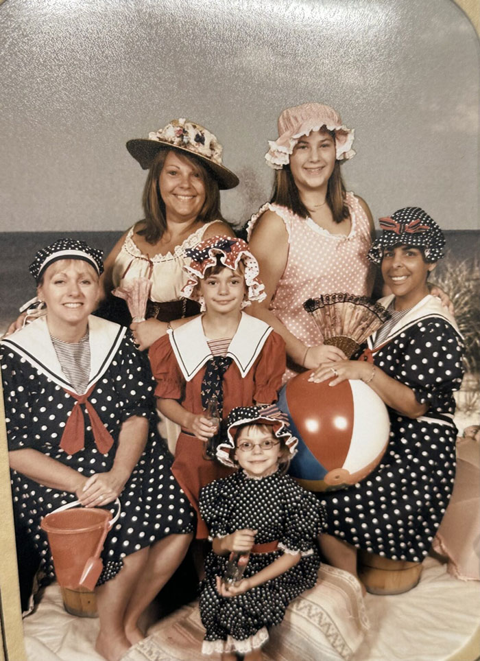 Group of women and children in vintage beach costumes posing with props in a funny vacation pic from a comedy special series.