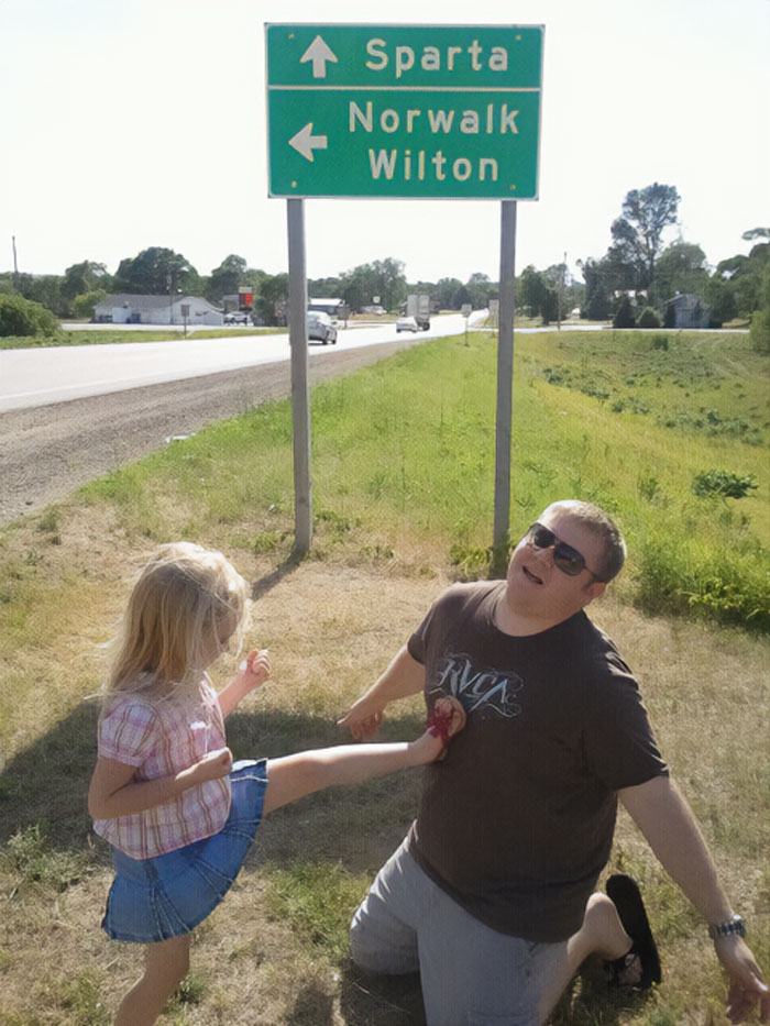 Young girl kicking man near road sign on grassy roadside, a funny moment from vacation pics that belong in a comedy special.
