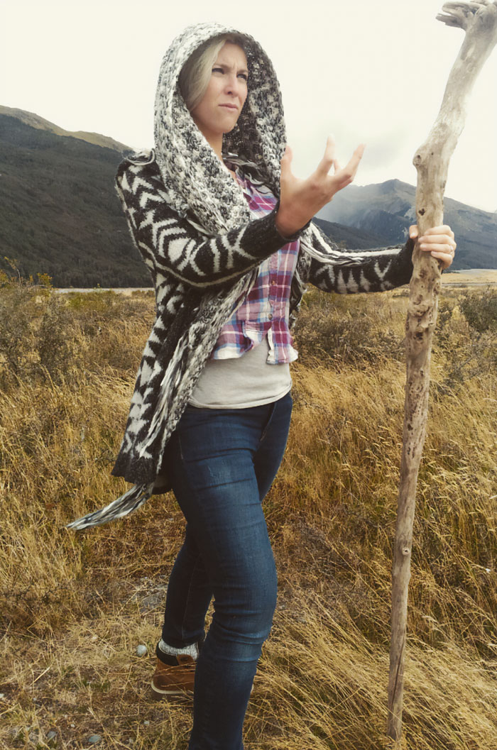 Woman in a patterned hooded sweater holding a walking stick in a field, a funny vacation pic with a comedy vibe.