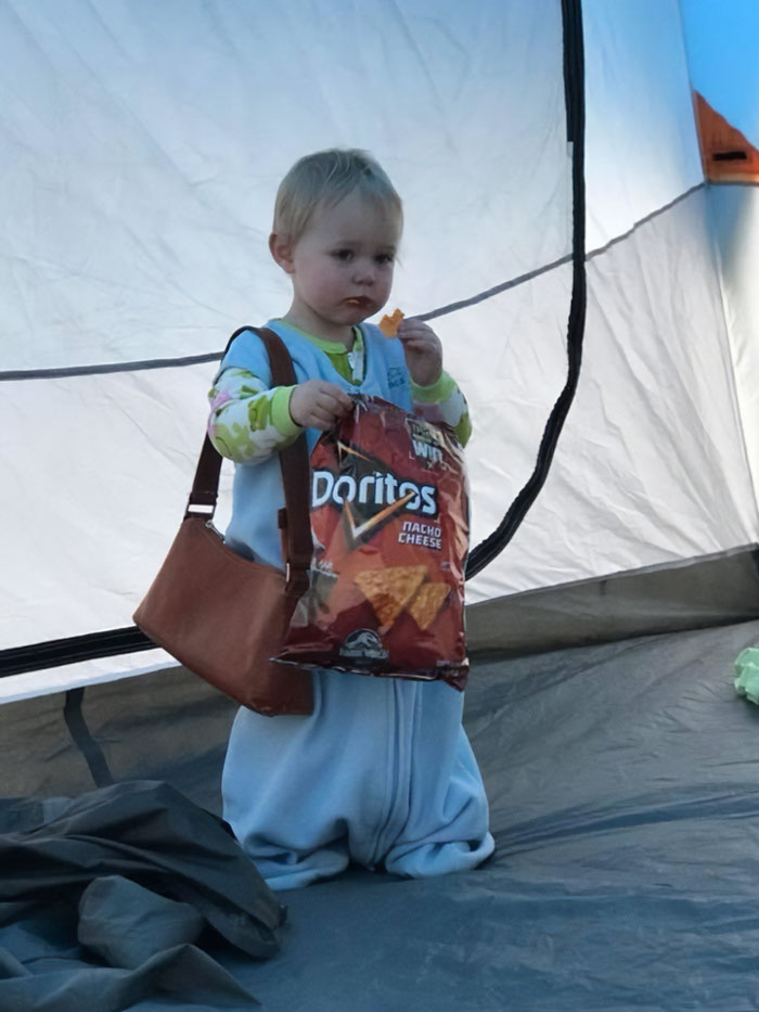 Toddler wearing a purse eating chips inside a tent, a funny vacation pic that looks like a comedy special moment.