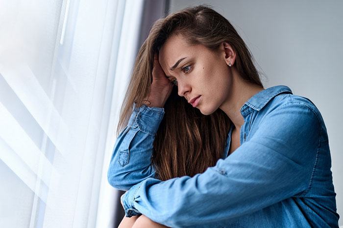 Young woman in a denim shirt looking worried by the window, reflecting on her genetic condition and pregnant sister. Young woman in a denim shirt looking worried by the window, reflecting on her genetic condition and pregnant sister.