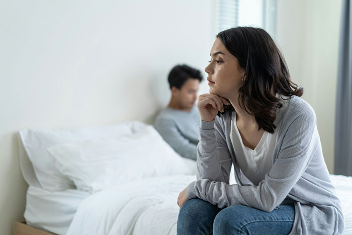 Woman sitting on bed looking thoughtful after breakup while man sits in background, dog updates implied. Woman sitting on bed looking thoughtful after breakup while man sits in background, dog updates implied.