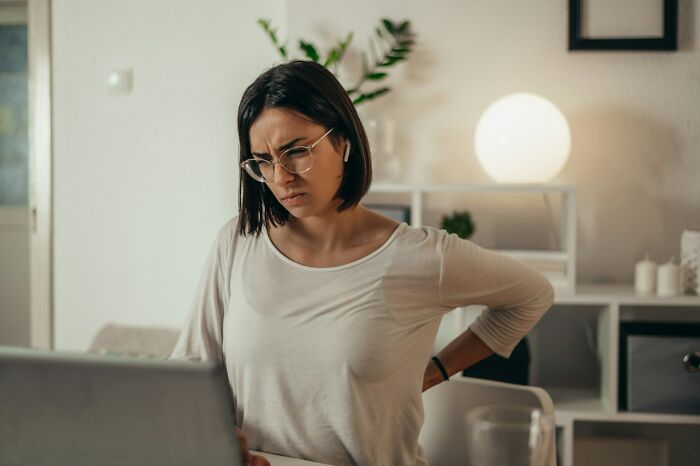 Young woman in glasses sits at a desk, holding her back in discomfort while looking at a laptop screen.