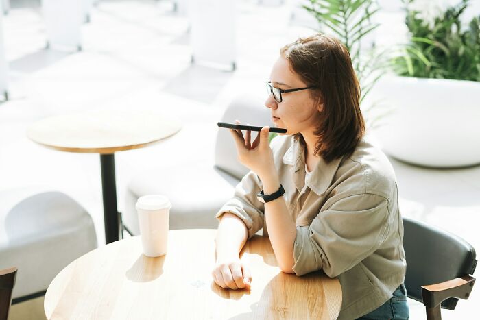 Young woman using voice recording on smartphone, representing glitches in the matrix people experienced concept in a bright cafe setting.