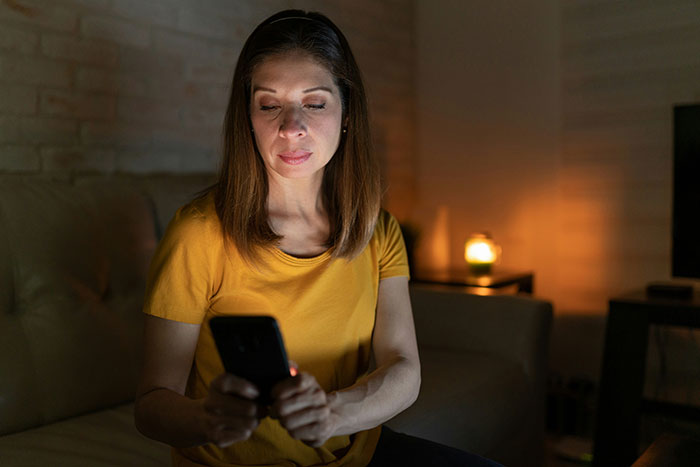 Mom-to-be reading strange text about fianc&eacute; on phone, sitting in dimly lit room, looking concerned and thoughtful.