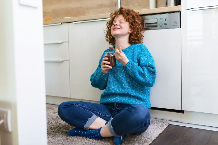 Young woman in a blue sweater enjoying a heartwarming moment while sitting on the kitchen floor with a jar of chocolate spread.