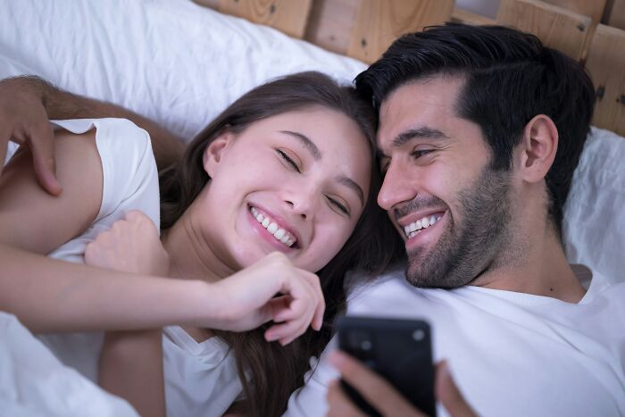A smiling couple lying in bed together, enjoying a moment while looking at a phone, showcasing smooth pickup lines.