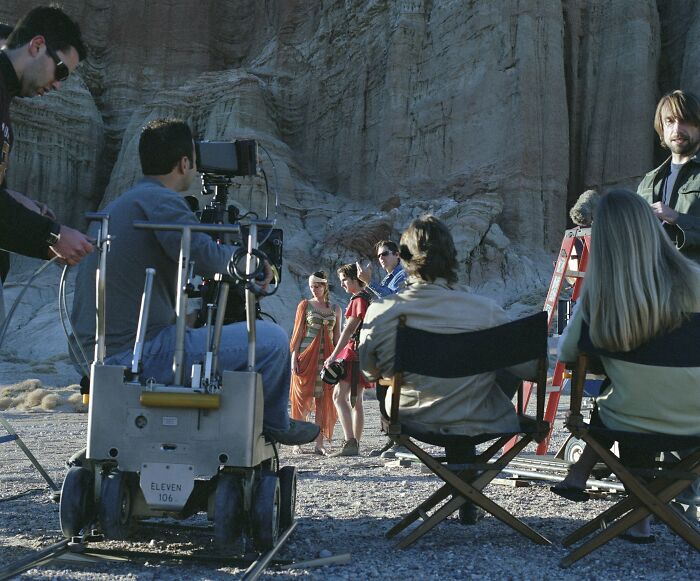 Film crew filming actors outdoors with rocky cliffs in the background, highlighting aspects of modern life breaking down.