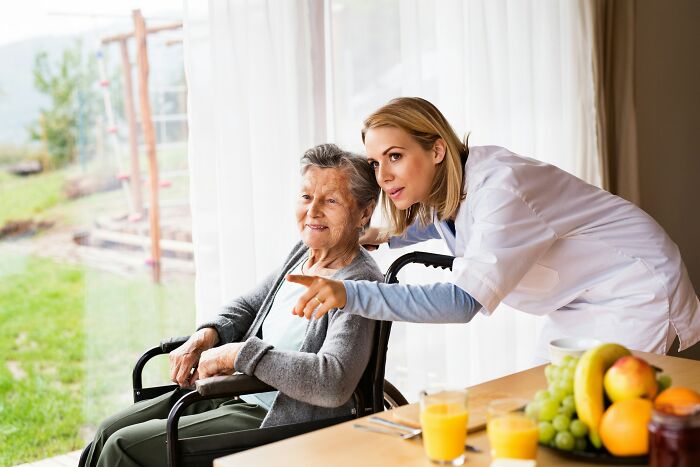 Caregiver in white coat pointing outside to elderly woman in wheelchair, illustrating aspects of modern life close to breaking down.