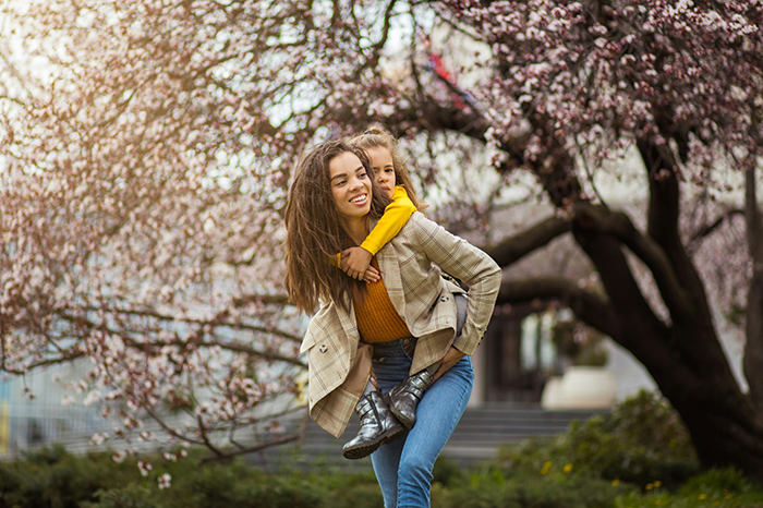 Smiling woman giving piggyback ride to child near blooming trees, capturing a joyful family moment outside.