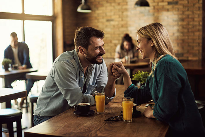 Couple laughing and holding hands at a cafe table, capturing GF confusion after BF&rsquo;s sis hitting on her situation.