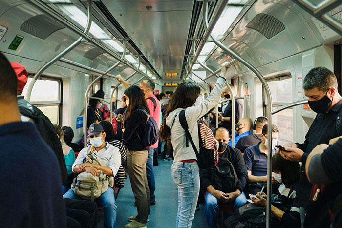 Young woman in a crowded subway car, standing and holding a handrail, people around wearing masks during commute.