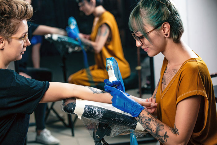 Tattoo artist with green hair and glasses applying a massive tattoo on a female client's arm in a studio.