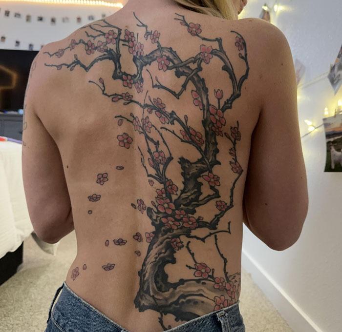Woman's back featuring a massive tattoo of a blossoming tree with pink flowers, highlighting girlfriend got massive tattoo.