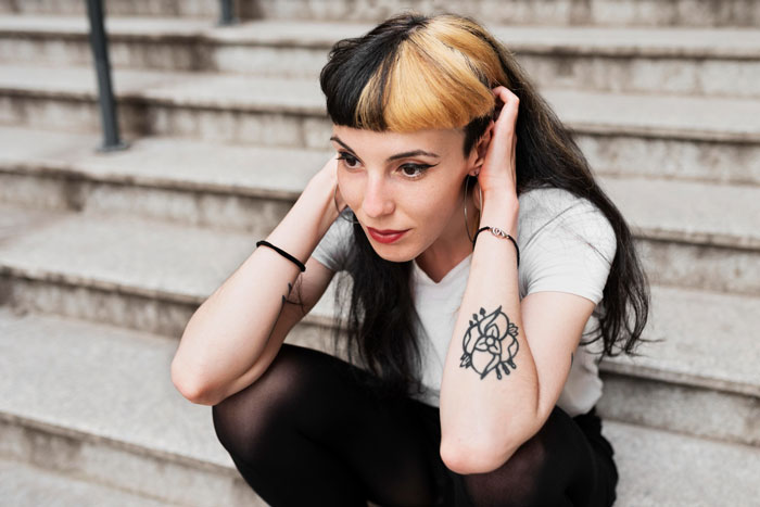 Young woman sitting on stairs showing massive tattoo on her arm with contrasting hair colors and hoop earrings