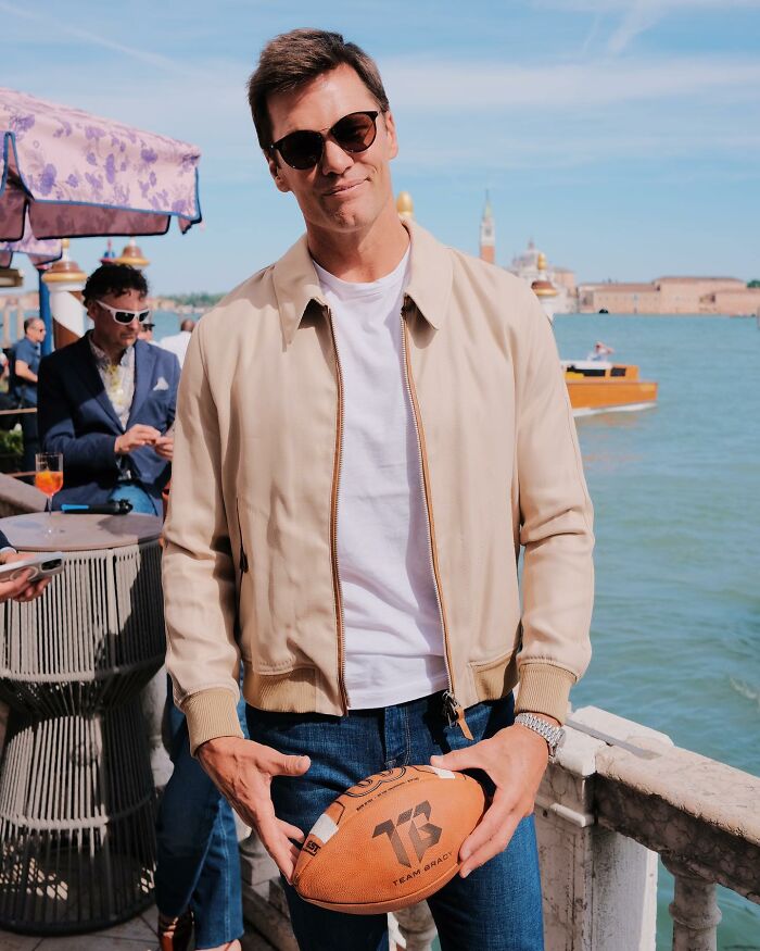 Tom Brady standing outdoors by the water holding a football, wearing sunglasses and a beige jacket.