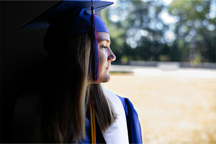 Graduate in cap and gown looking thoughtful and crushed while gazing out a window on graduation day.