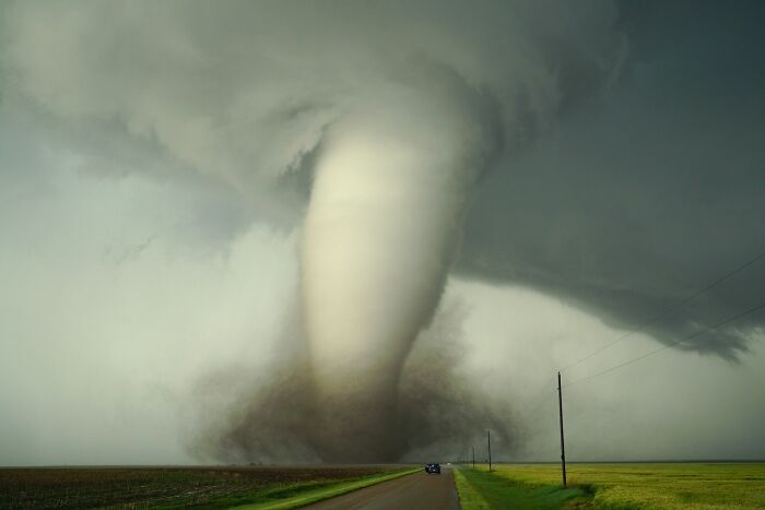 Massive tornado touching down on open farmland with a car driving away, representing people surviving crazy situations.