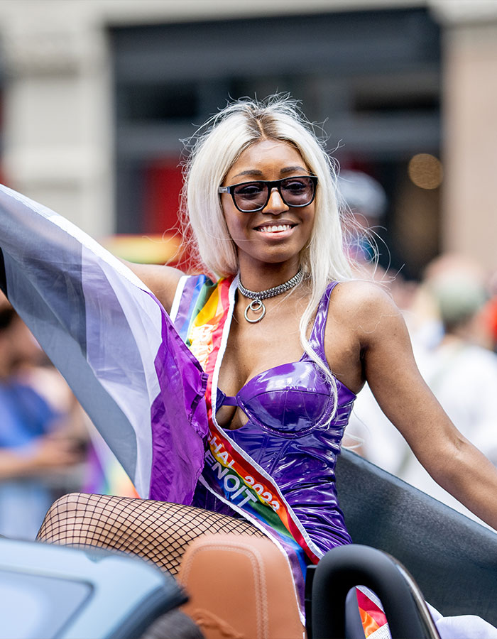 Person wearing a purple outfit and sunglasses holding a flag representing greys*xuality at a modern dating culture event.