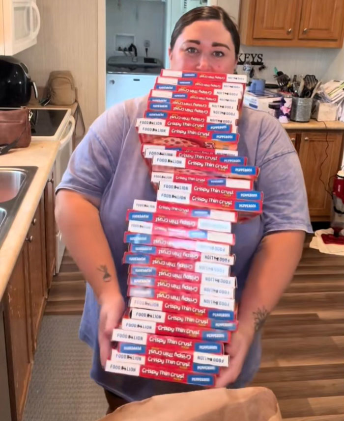 Woman in kitchen holding a large stack of frozen pizzas as part of an American family grocery haul