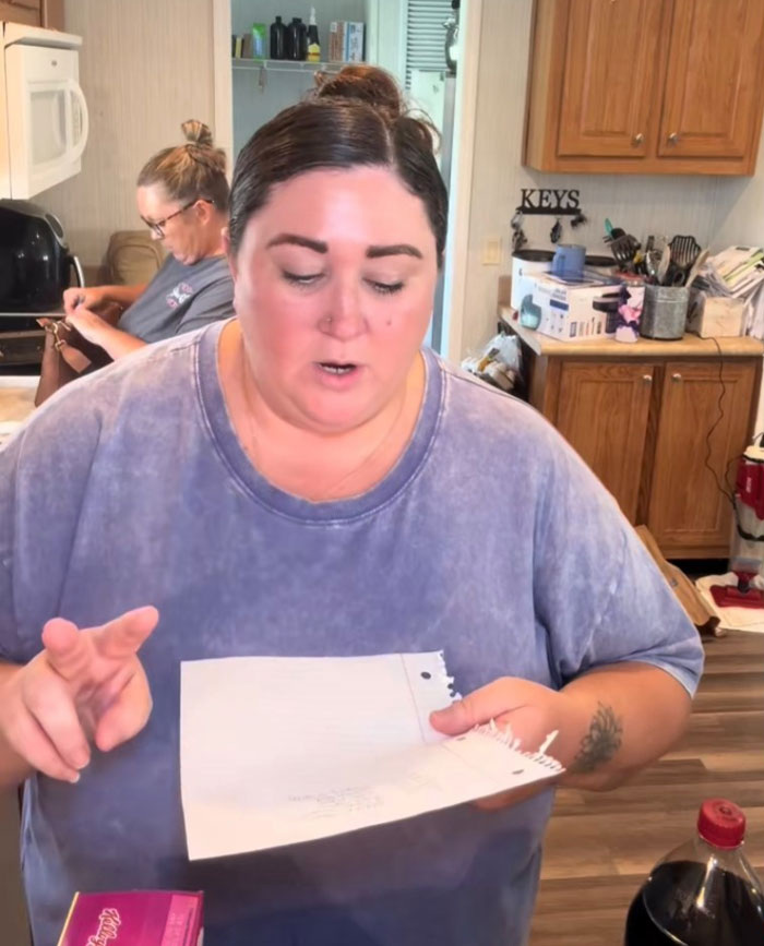 Woman in a kitchen reviewing a grocery list as part of an American family grocery haul with cereal and soda nearby.