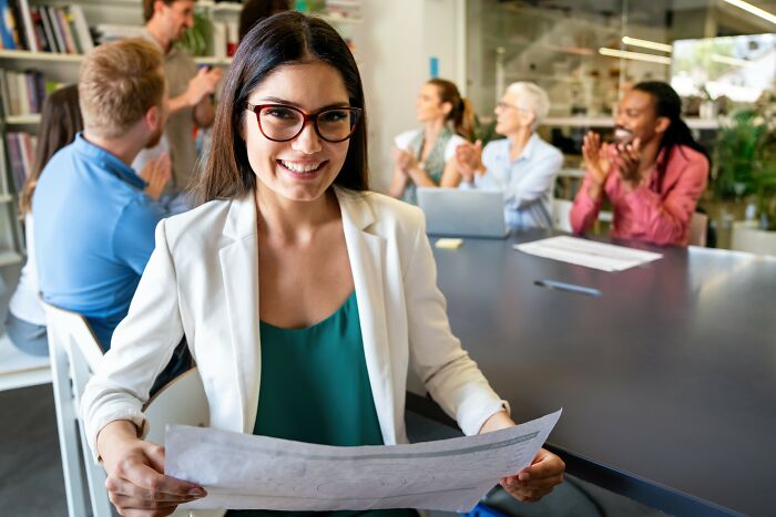 Young professional woman smiling confidently holding documents while colleagues discuss and applaud in modern office setting, smartest kids