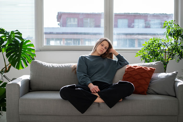 Young woman sitting on a gray sofa looking pensive, representing reluctance as future caregiver for autistic stepbrother.