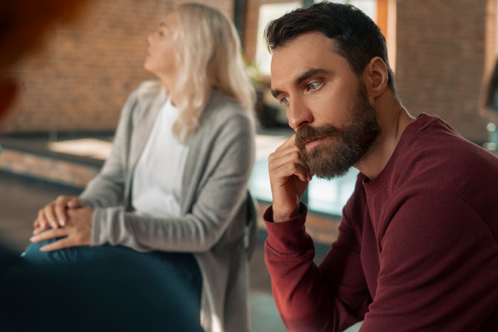 Young man looking thoughtful while woman in background avoids eye contact, representing autistic stepbro and caregiver conflict.