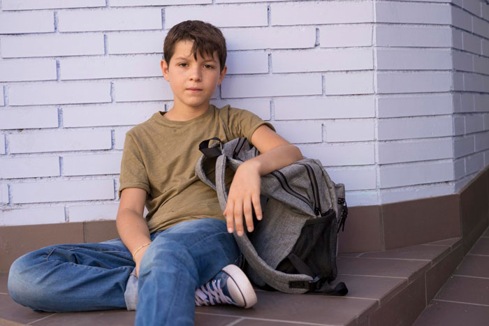 Young boy sitting on steps with backpack, looking thoughtful and upset, reflecting feelings of neglect in family conflict.