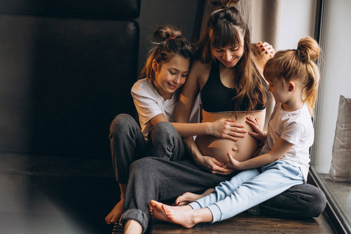 Pregnant lady sitting with her children, smiling and touching her baby bump in a warm family moment at home.