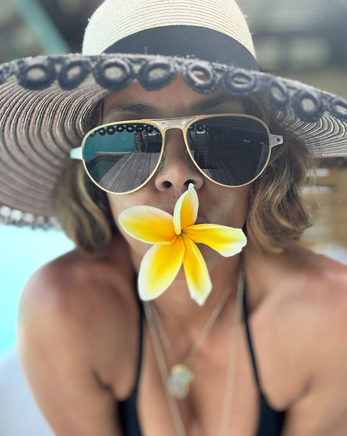 Close-up of a woman wearing sunglasses and a wide-brimmed hat with a yellow flower in her mouth, reflecting a wooden deck.