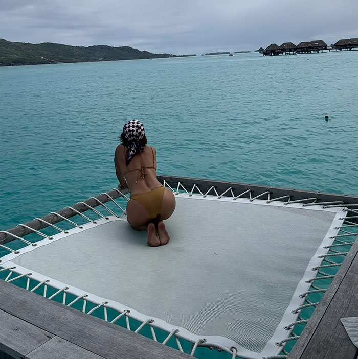 Woman in bikini kneeling on water hammock overlooking ocean, related to Halle Berry trolls ex-husband cooking and cleaning claims