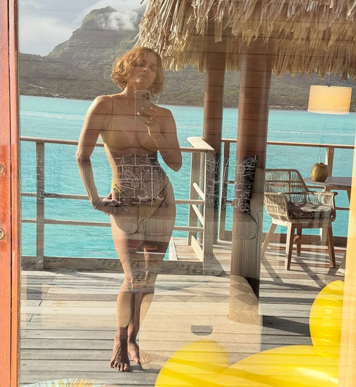 Halle Berry taking a mirror selfie in swimwear with ocean and mountains in the background, trolling ex-husband cooking cleaning claims.