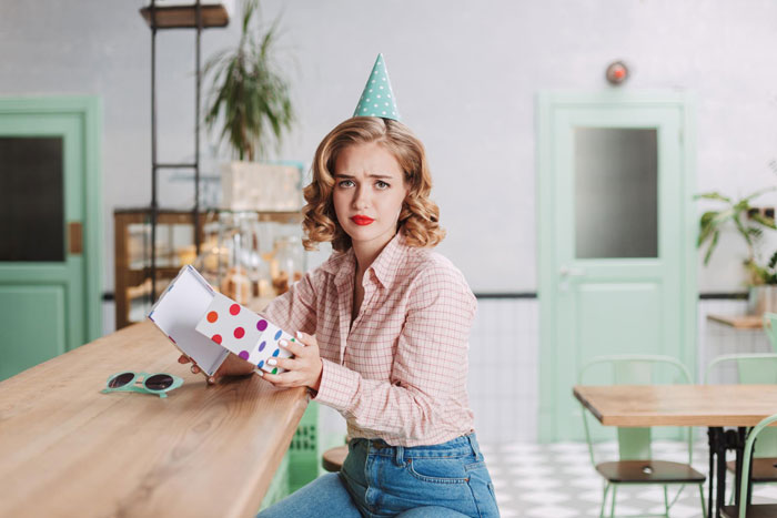 Young woman wearing a party hat at a birthday dinner, upset while holding a gift box, influencer filming conflict scene.