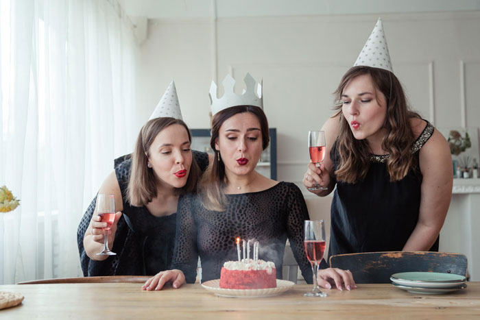 Three women at a birthday dinner, one wearing a paper crown, with party hats and glasses of pink drink celebrating.