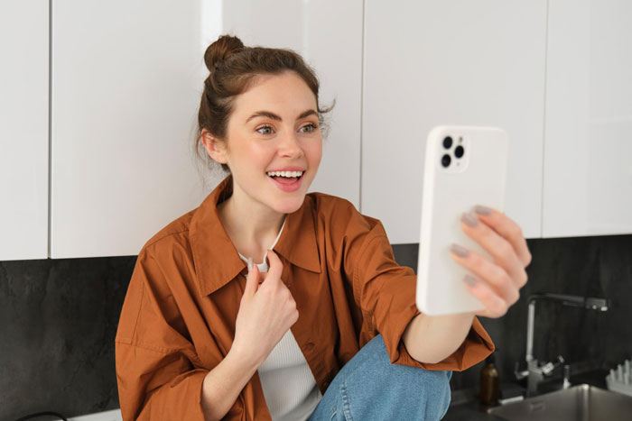 Young woman acting as self-proclaimed influencer filming herself with smartphone, looking excited in casual indoor setting.