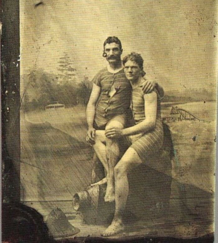 Two handsome Victorian men in vintage swimwear posing together by the water in an old sepia photograph.